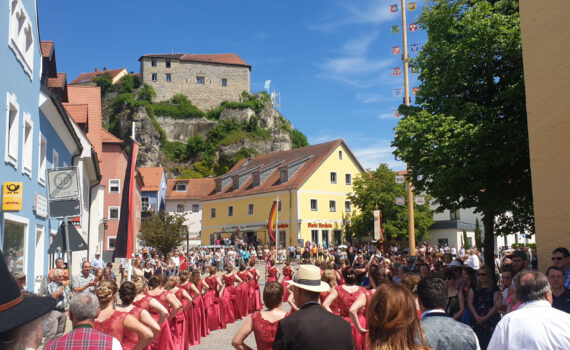 Festzug am Markt Laaber. Links und rechts Gebäude. Im Hintergrund Blauer Himmel und ein Grün bewachsener Berg mit Burgruine