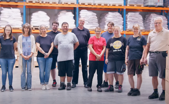 11 Personen stehen Lächelnd nebeneinander in einer Lagerhalle. Das Team von Purux
