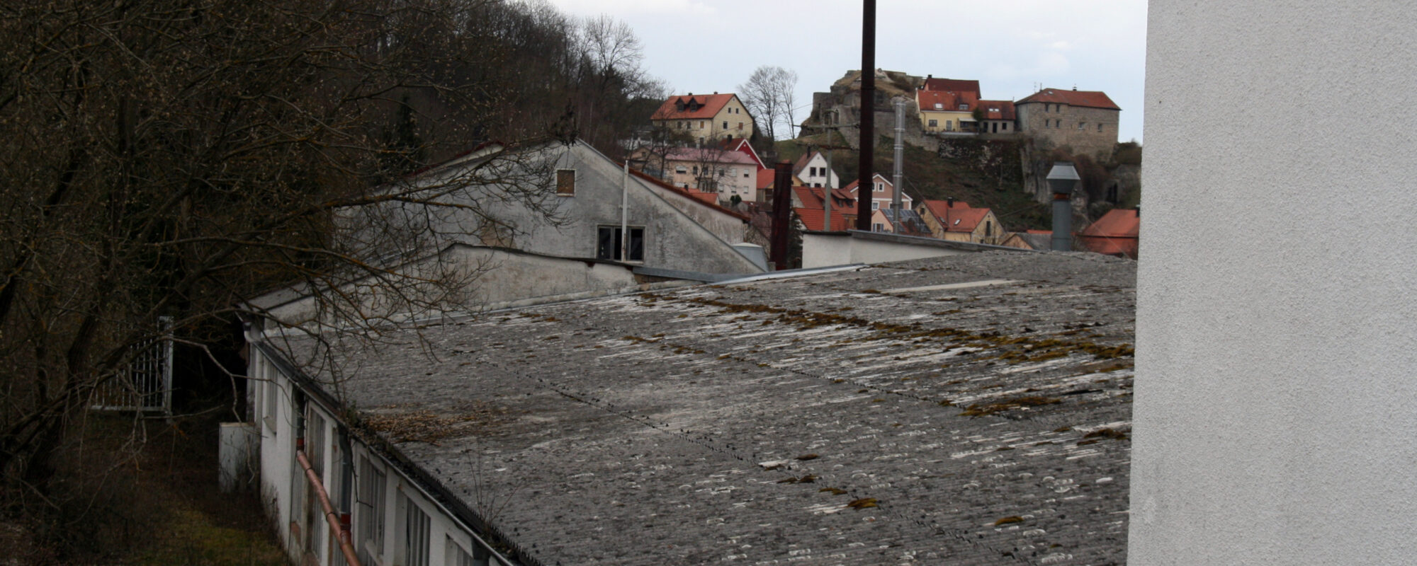 Die alte ehemalige Halle. Sehr heruntergekommen und niedrig gebaut. Links ein paar trockene Bäume. rechts im Hintergrund Die Burgruine und Häuser.