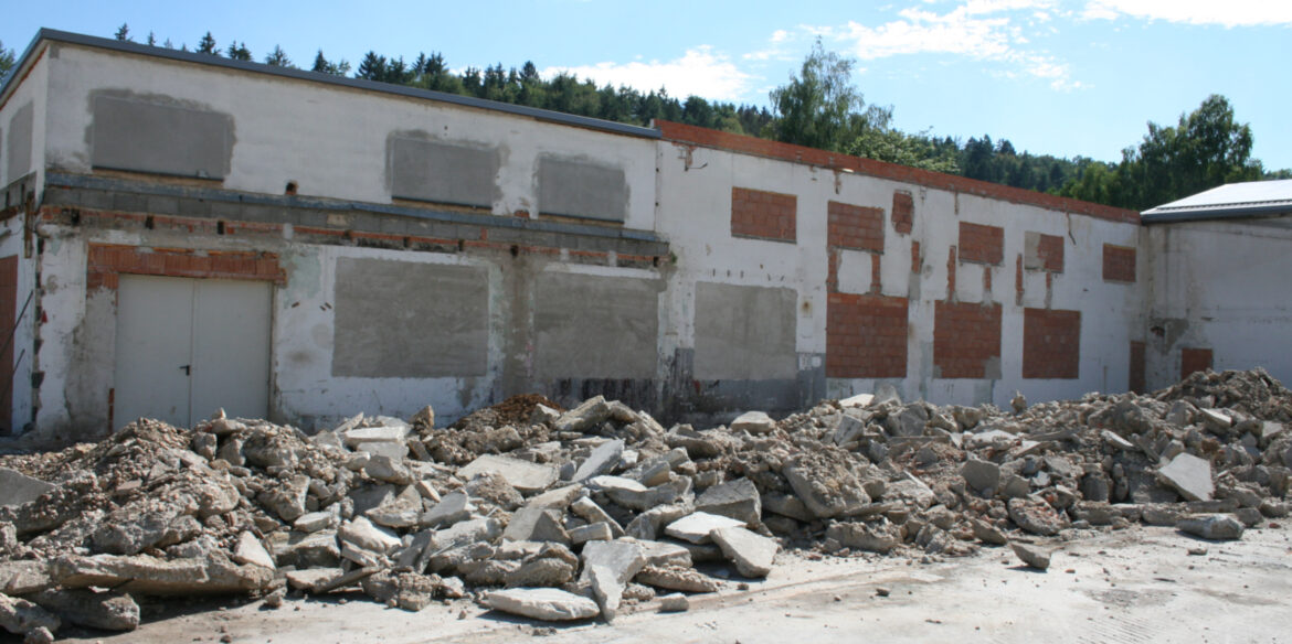 Eine Kaputte Mauer mit einer Großen Tür auf der linken Seite. Vor der Mauer liegen viele Beton stücke.
