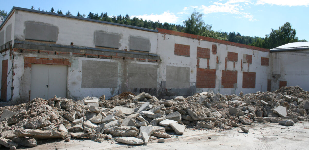 Eine Kaputte Mauer mit einer Großen Tür auf der linken Seite. Vor der Mauer liegen viele Beton stücke.