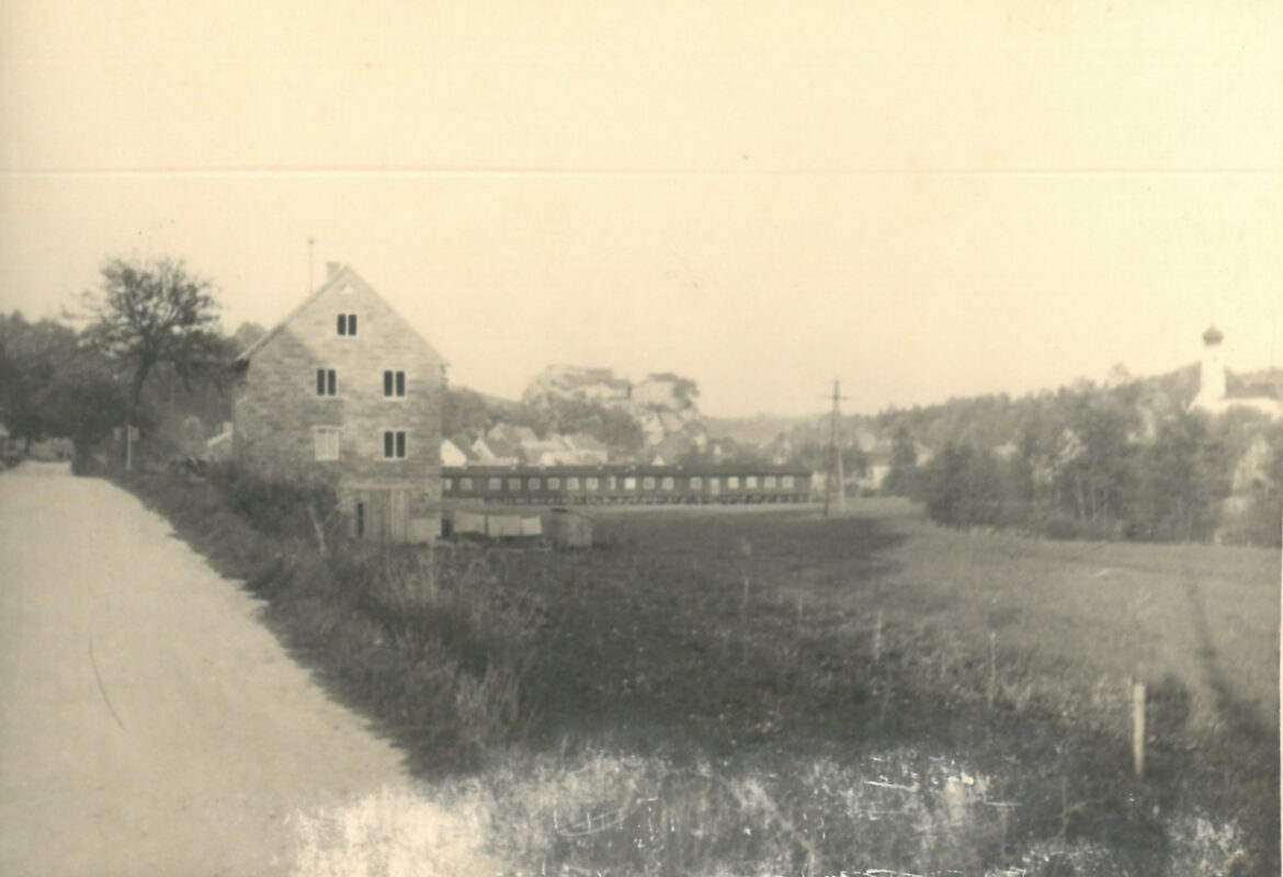 Altes schwarz weiß Bild. Ehemalige Firma damals bestehend aus einem Gebäude und einer langen brücke. Links neben der Firma entlang führt eine Schotter Straße zwischen ein paar bäumen hindurch. Rechts stehen ein paar Bäume und eine Kirche. Im Hintergrund ist die Burgruine auf einem Berg und der Markt Laaber bestehend aus einer Ansammlung von Häusern.