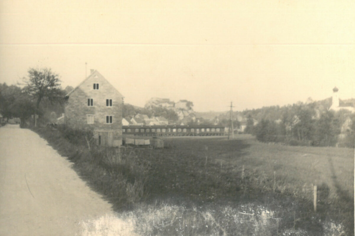 Altes schwarz weiß Bild. Ehemalige Firma damals bestehend aus einem Gebäude und einer langen brücke. Links neben der Firma entlang führt eine Schotter Straße zwischen ein paar bäumen hindurch. Rechts stehen ein paar Bäume und eine Kirche. Im Hintergrund ist die Burgruine auf einem Berg und der Markt Laaber bestehend aus einer Ansammlung von Häusern.
