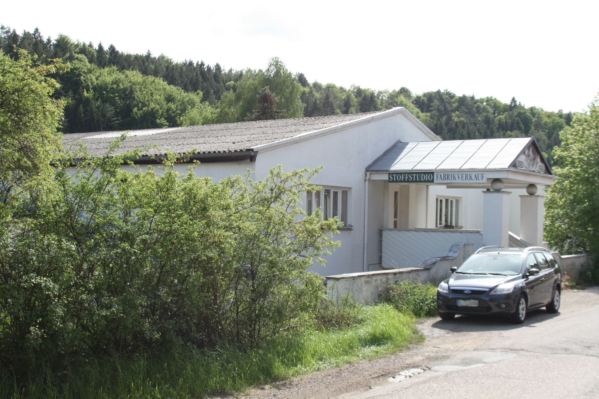 Altes Bild des Firmeneingangs. Überdachte Treppen mit mauern links und rechts. An dem Dach der Treppen steht Stoffstudio Fabrikverkauf geschrieben
