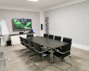 Weiß gehaltener raum mit großem Bildschirm an der Wand. Langer schwarzer Holztisch umgeben von Schwarzen Stühlen in der Mitte.